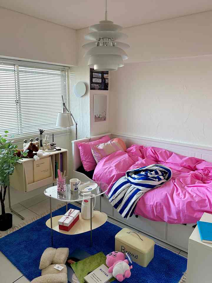 白い壁とベッドフレームに明るいピンクの寝具が置かれた小さな寝室、青いラグと自然光が調和した居心地の良い空間