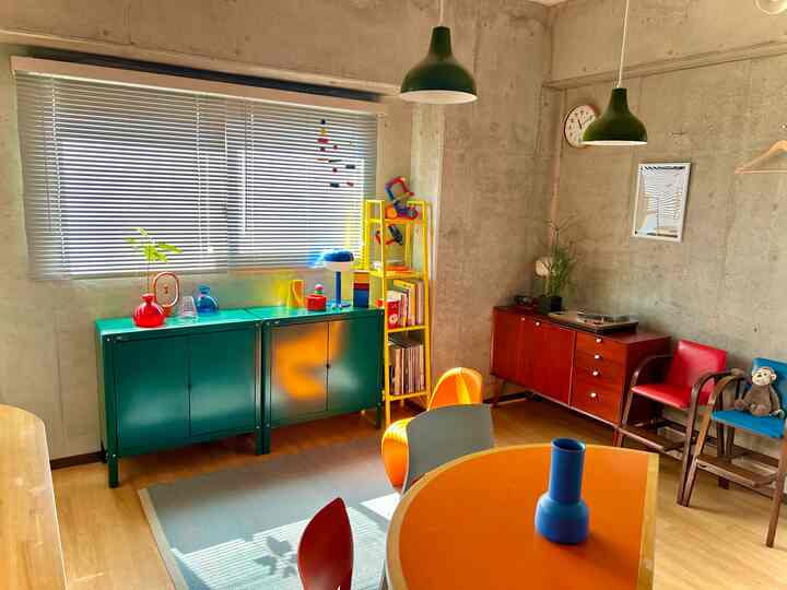 ナチュラルな木製床とコンクリート壁のミッドセンチュリーモダンなリビング、カラフルなキャビネットと椅子が配置された印象的な空間