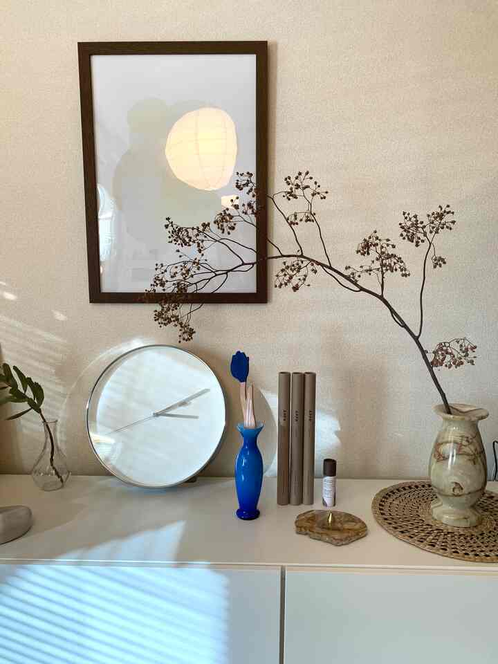 白い壁と家具の上にアートフレーム、白い時計、青い花瓶、自然素材の大理石の花瓶と乾燥した枝が配置されたシンプルな空間