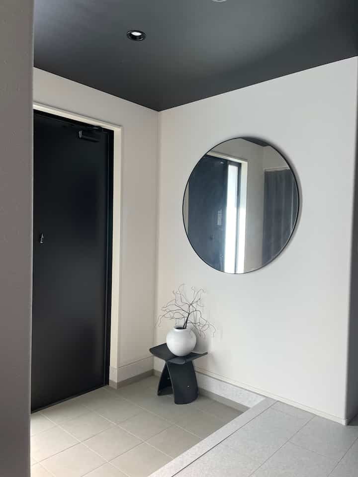 白い壁と黒い玄関ドア、円形の鏡と黒いスツールが配置されたシンプルでモダンな玄関スペース