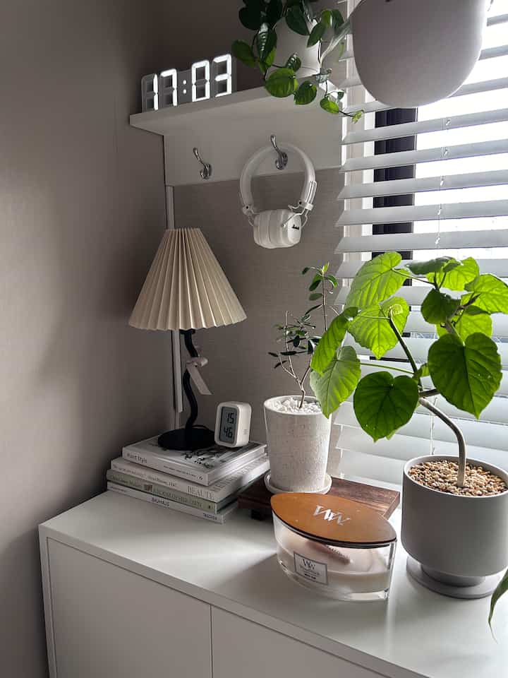 ホワイトとベージュのトーンの小さな空間、デジタル時計、テーブルランプ、アロマキャンドル、緑の植物が調和したシンプルなインテリア