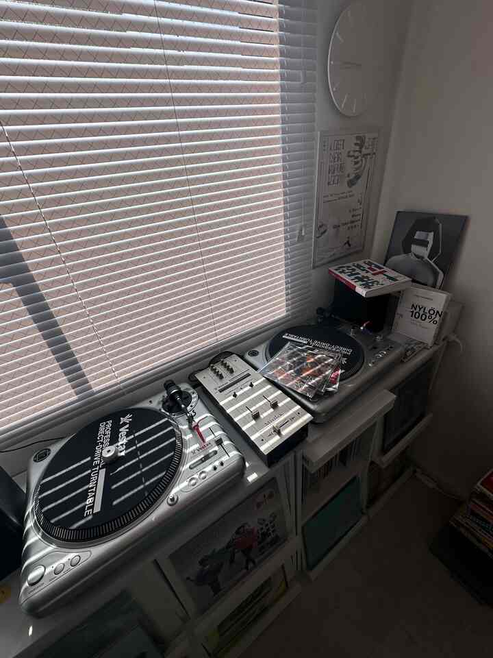 ホワイトトーンのコンパクトな部屋、ブラインドの窓際にディスプレイラックとヴィンテージのターンテーブルが配置されたレトロな雰囲気の空間