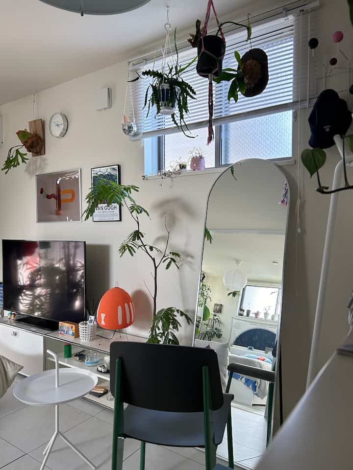 明るいホワイトトーンのリビング、様々な緑の植物と大きな全身鏡、モダンなサイドテーブルとテレビ台が配置された雰囲気のある空間