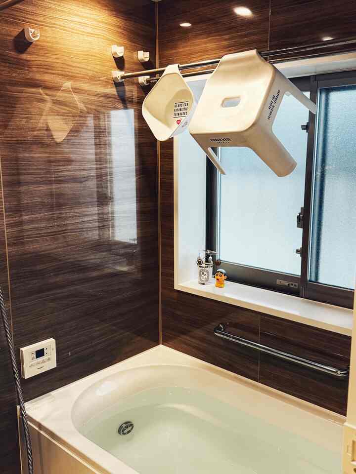 ナチュラルカラーのバスルーム、窓際にヒューマンメイドのバスチェアとシャワーペールが吊るされている雰囲気のあるインテリア