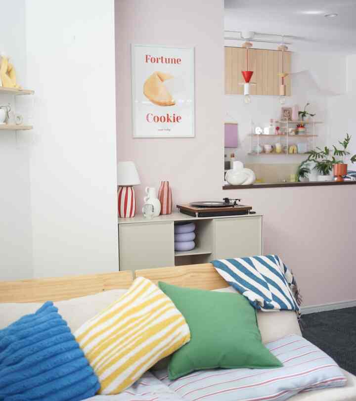 柔らかなピンクと白の壁のリビング、木製フレームのソファとカラフルなクッション、キャビネット上のポスターとレトロなレコードプレーヤーが配置された雰囲気のあるインテリア