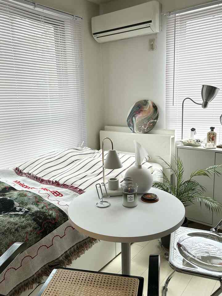 White-toned compact bedroom featuring a round table, bed, window blinds, and floor lamp creating a simple modern space