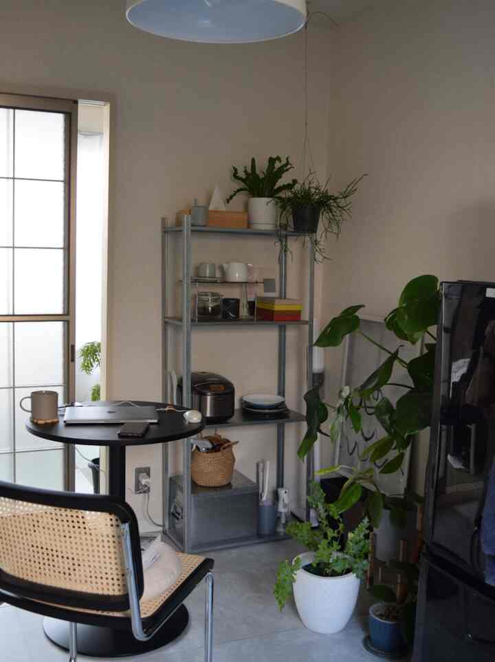 ライトグレーのタイル床と白い壁の小さなダイニングルーム、モダンな丸テーブルと椅子、植物とコーヒー用品が置かれた金属製の棚が配された雰囲気のあるインテリア