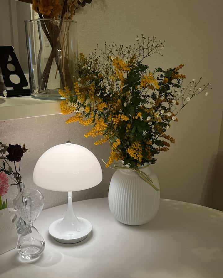 白を基調としたテーブルに、明るいテーブルランプと黄色いミモザの花が生けられた白い花瓶が配置された雰囲気のあるインテリア