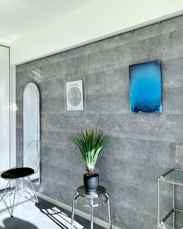 グレー調の無機質な壁面と人工観葉植物が置かれたミニマルな空間、スチール製スツールとワイヤーチェアが配置された印象的な玄関インテリア