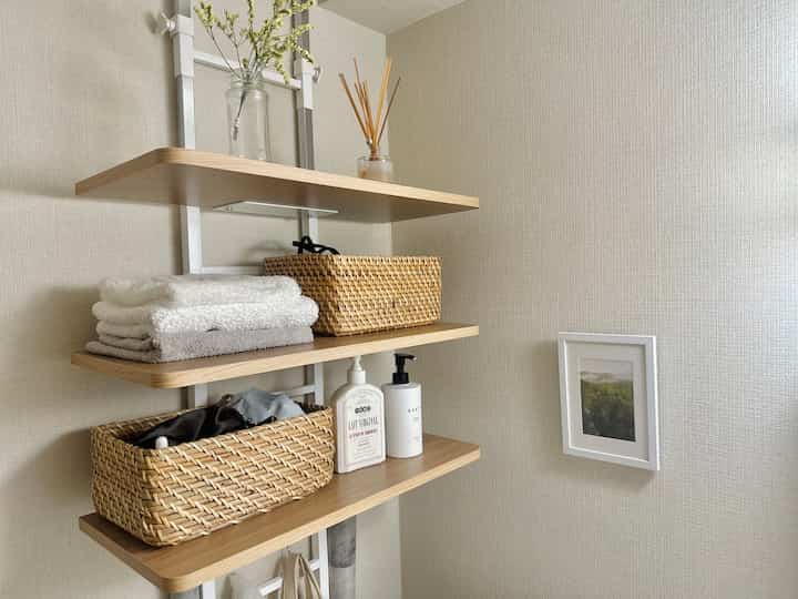 ベージュ色の壁と木目調の棚があるバスルーム空間、ラタンのバスケットとタオル、ディフューザーが整った印象的なインテリア