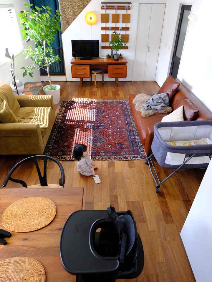 温かみのある木目調とナチュラルな色合いのリビング、ブラウンのソファとグリーンのアームチェア、ベビーベッドとラグが配置された赤ちゃんと暮らす空間