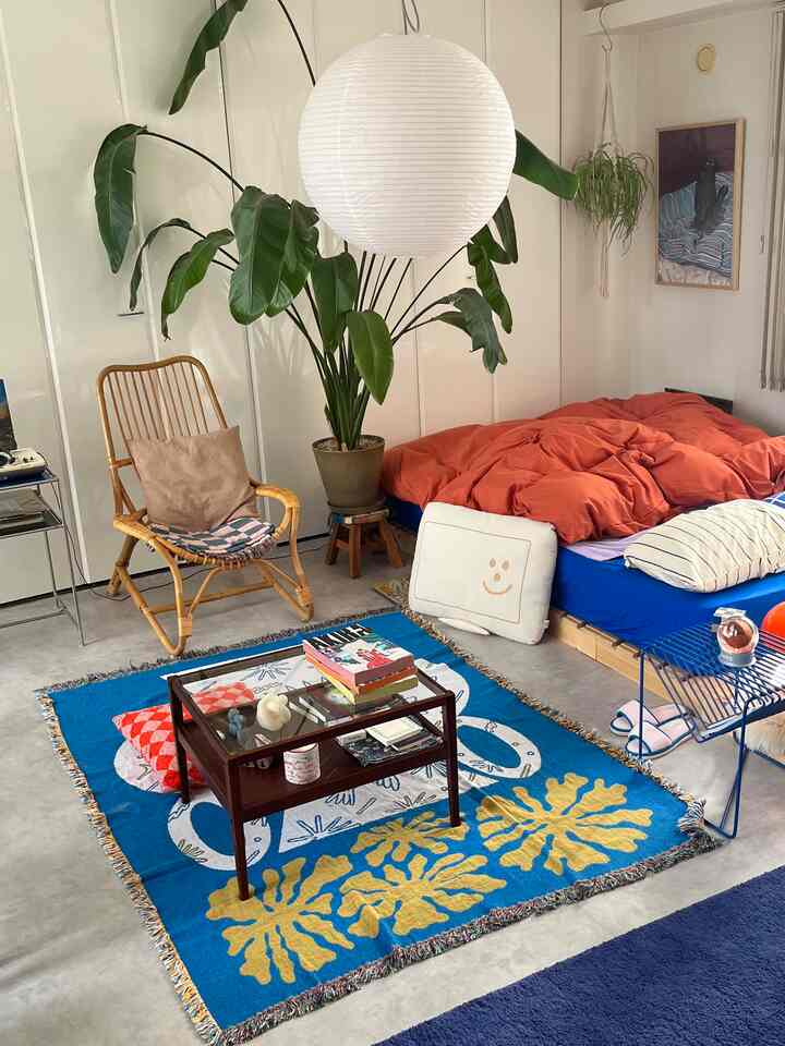 Blue rug covered bedroom featuring orange bedding, rattan armchair, and large potted plant with a natural vibe