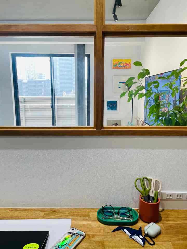 白い壁と木目調の窓枠が印象的なワークスペース、机の上にはトレイやはさみが配置された落ち着いた雰囲気のインテリア