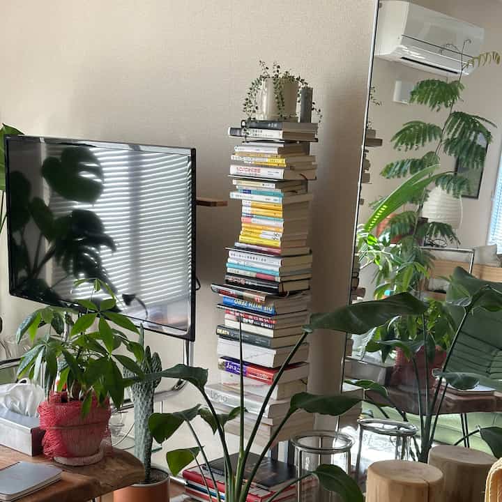 白い壁のリビング、豊富な緑の植物、大きな本のタワーと木製テーブルが配置された自然な雰囲気のインテリア