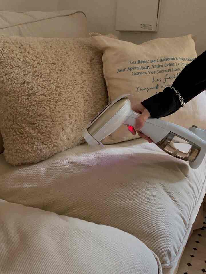 暖かいベージュのトーンのリビングのソファとクッションの上で、手が白い衣類スチーマーを持っている様子