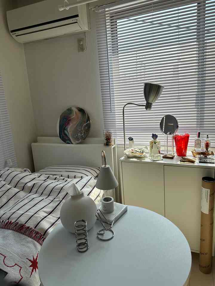 白とグレーの色調のコンパクトな寝室、シンプルなベッドと丸テーブル、シルバーの照明が配置された清潔感のある空間