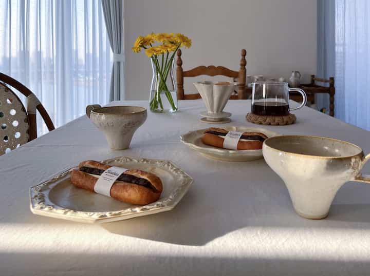 白を基調としたキッチンダイニングテーブル、黄色い花の花瓶、陶器のマグカップ、コーヒーポット、パンが配置された雰囲気のあるおうちカフェ空間