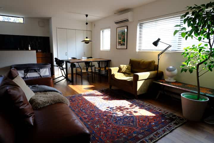 白い壁と天井のリビング、茶色のレザーソファとマスタード色のアームチェア、ペルシャ風ラグが配置された自然でモダンな雰囲気の空間