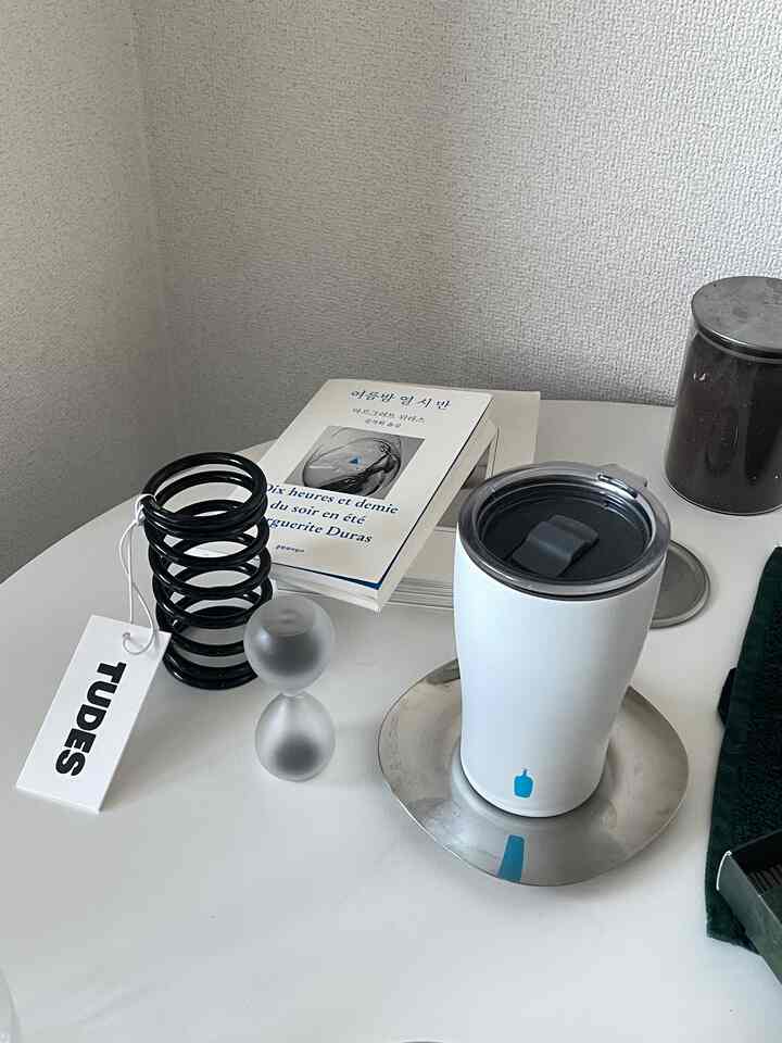 A clean home cafe setup on a monotone white table featuring TUDES stainless steel holder, hourglass, and Blue Bottle coffee cup
