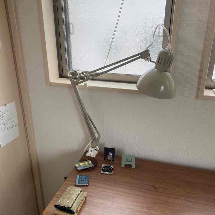 白い壁と窓があり、木目調のデスクの上に大きな白い作業用ランプとデジタルアラーム時計が置かれたコンパクトなホームオフィス空間