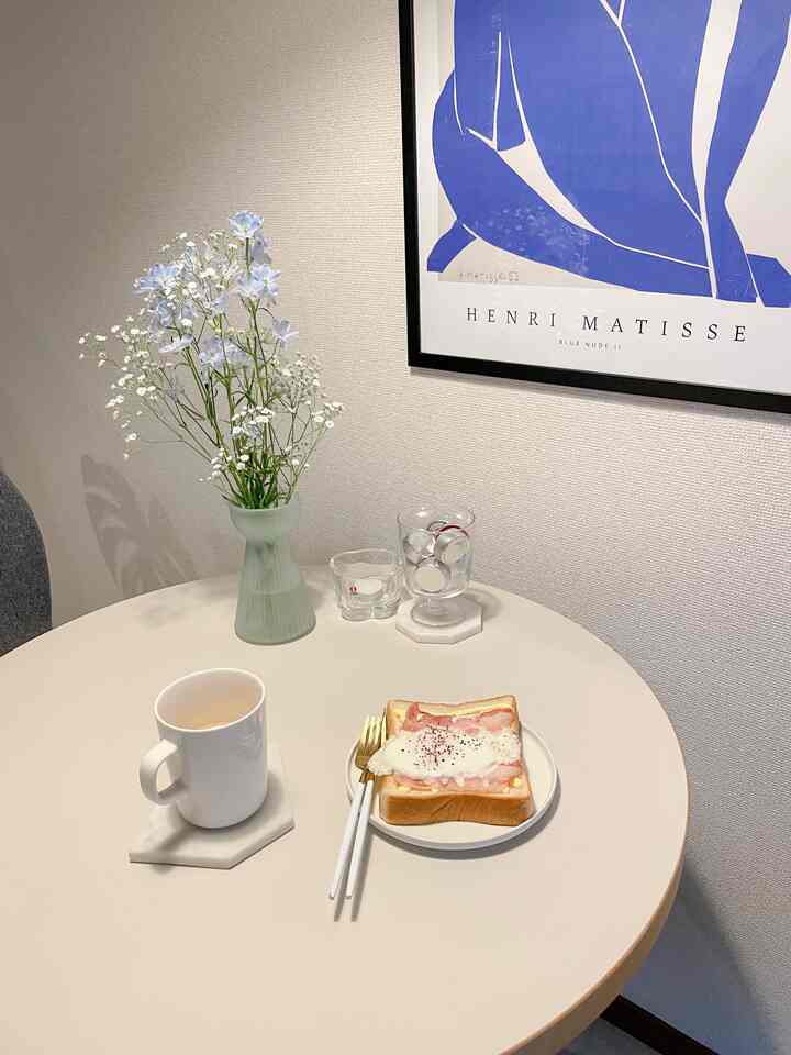 ホワイトとブルーの色調のダイニング、丸テーブルにマグカップとベーコントースト、花瓶を配置した雰囲気のある朝の空間