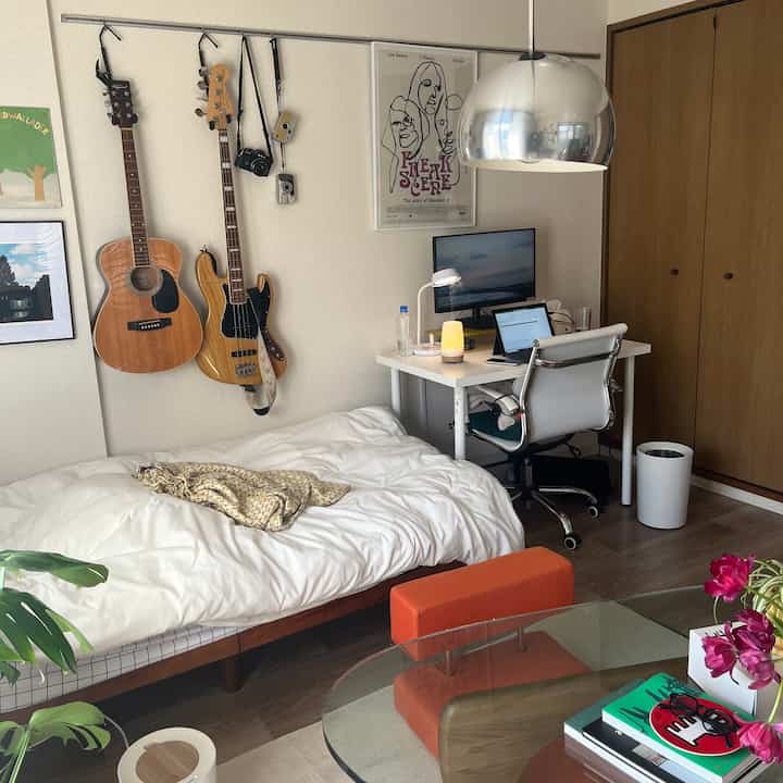 白と茶色のトーンが特徴的な小さな寝室兼ホームオフィス、壁にギターが掛けられた一人暮らし向けの落ち着いた空間