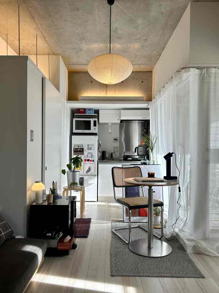 White and natural toned studio apartment featuring a compact kitchen, small round dining table, and pendant lighting creating a cozy atmosphere