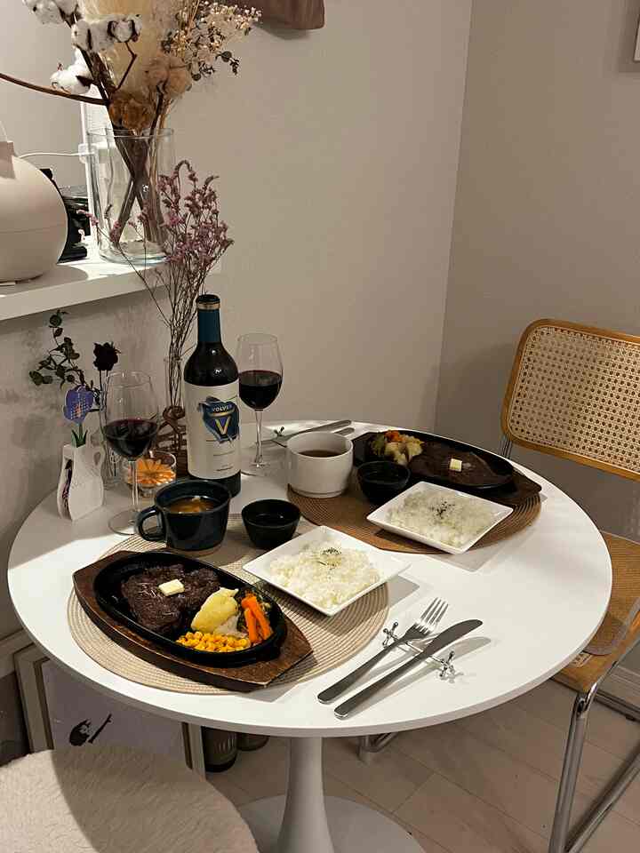 白と木目調の調和が感じられるダイニング、シンプルな丸テーブルにステーキの料理とワイングラス、花瓶が配置された雰囲気のあるおうちデート空間