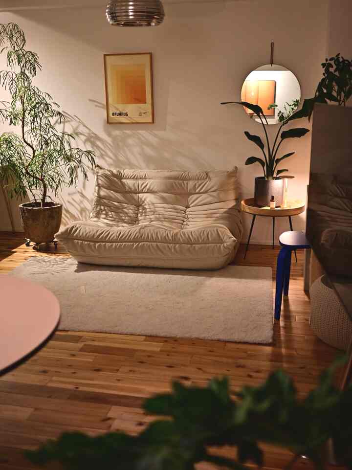 ナチュラルな木目調と白を基調としたリビング、クリーム色のソファと丸い鏡、植物が配置されたシンプルで温かみのある夜の雰囲気のインテリア