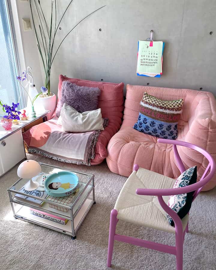 A pink-toned living room with concrete walls featuring two pink sofas and a pink-framed chair arranged in a cozy setting