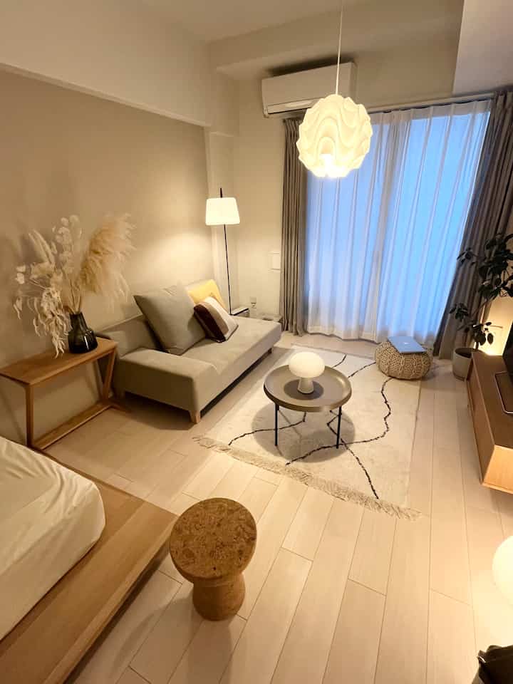 Natural beige-toned studio apartment bedroom and living room featuring modern sofa, pendant light, and cozy ambiance