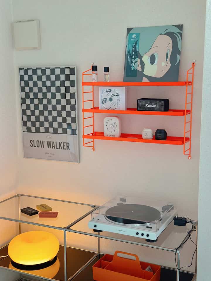 Minimal music corner with white walls, bright orange wall shelf, transparent stand holding white turntable, and checkerboard poster
