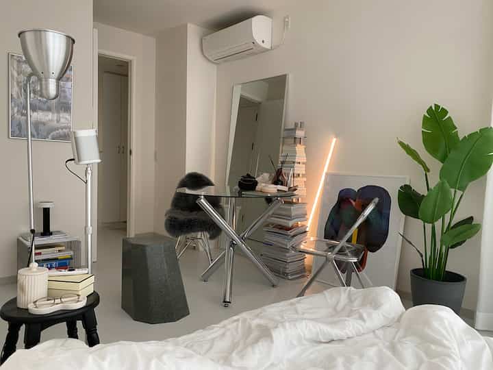 白とシルバーのトーンの寝室、ガラスのデスク、スツール、フロアランプ、緑の植物が配置されたミニマルで明るいインテリア