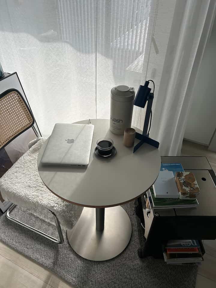 White and silver toned studio apartment dining space with a round table featuring a laptop and coffee items, conveying a modern atmosphere