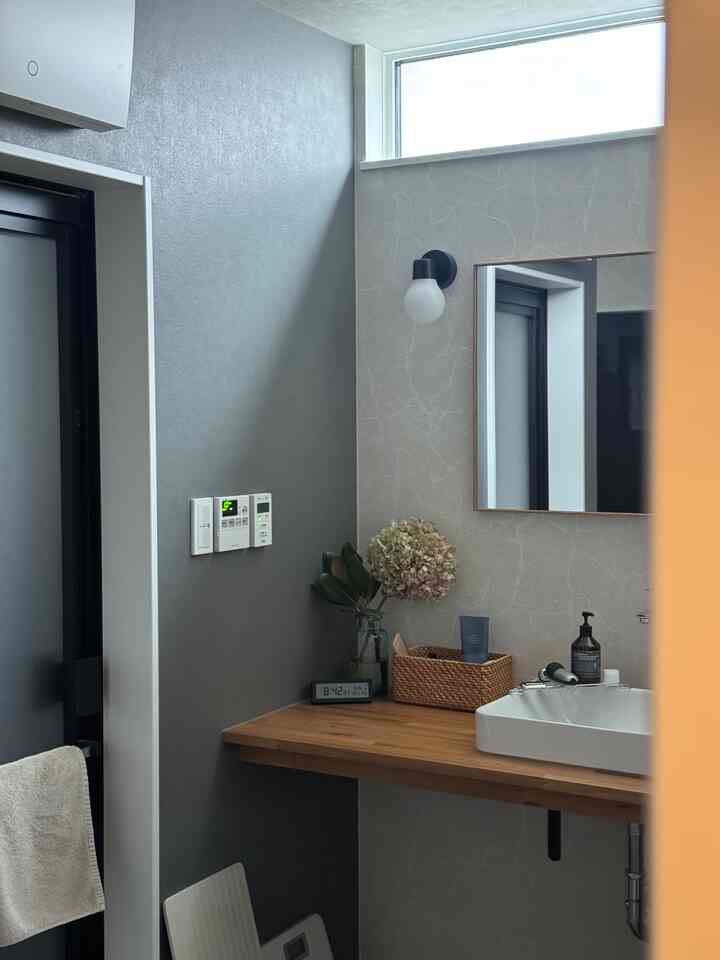 グレーと木目調の壁のあるトイレ空間、デジタル時計とラタンバスケットが置かれたナチュラルモダンな洗面台