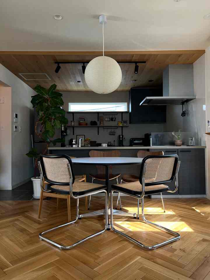 木目調の床と白い丸いダイニングテーブルが中心のダイニング、モダンなキッチンとペンダントライト、大きな観葉植物が配置された自然な雰囲気のインテリア