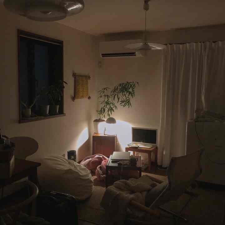 暖かくナチュラルな色調の小さな一人暮らし向け寝室、木製家具とテーブルランプ、白いカーテンとビーズクッションが配置された雰囲気のある空間