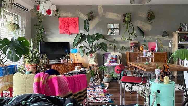 Dark brown floored natural-toned living room featuring colorful indoor plants, vintage furniture, and pink accents in a cluttered space