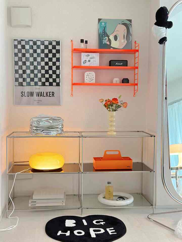 A white and orange toned space featuring transparent shelves, unique lighting, and a vase, reflecting a modern and minimal interior