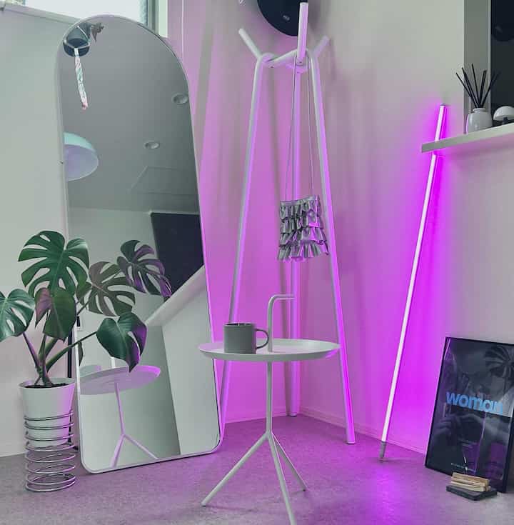 ホワイトとピンクの照明が印象的なリビングコーナー、サイドテーブルやコートラック、大型の鏡、植物スタンドが配置されたモダンなインテリア