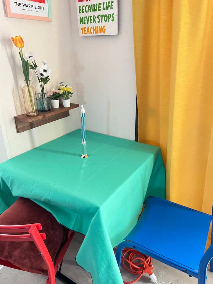 パステル調の色合いと黄色のカーテンが印象的なダイニング、ティール色のテーブルとカラフルな椅子が配置された雰囲気あるおうちカフェ空間