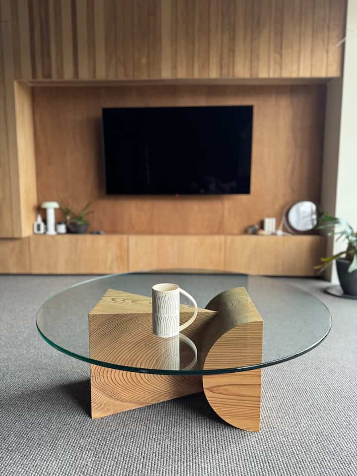 ナチュラルな木材とグレーのカーペットが調和したリビング、中央にガラス製丸テーブルとパターン入りのマグカップが配置された印象的な空間