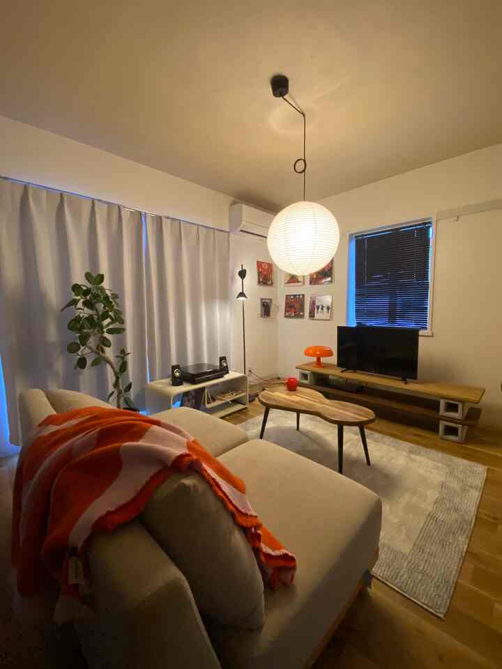 A white-walled, wood-toned 6-tatami living room featuring a beige sofa, mid-century modern pendant light, and vibrant orange mushroom lamp creating a cozy atmosphere
