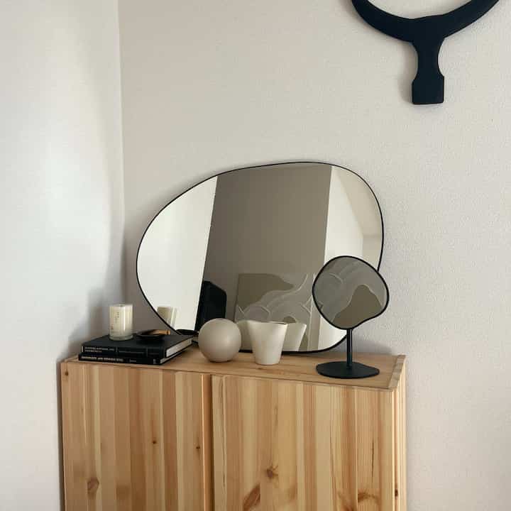 Simple studio apartment with natural wood tone cabinet topped with two irregular-shaped mirrors against white walls