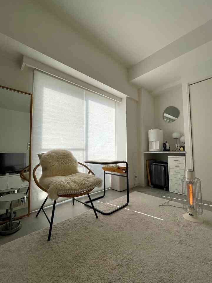 白とベージュの落ち着いた色調のコンパクトなリビング、アームチェアとサイドテーブル、ブラインドが配置された自然な雰囲気のインテリア