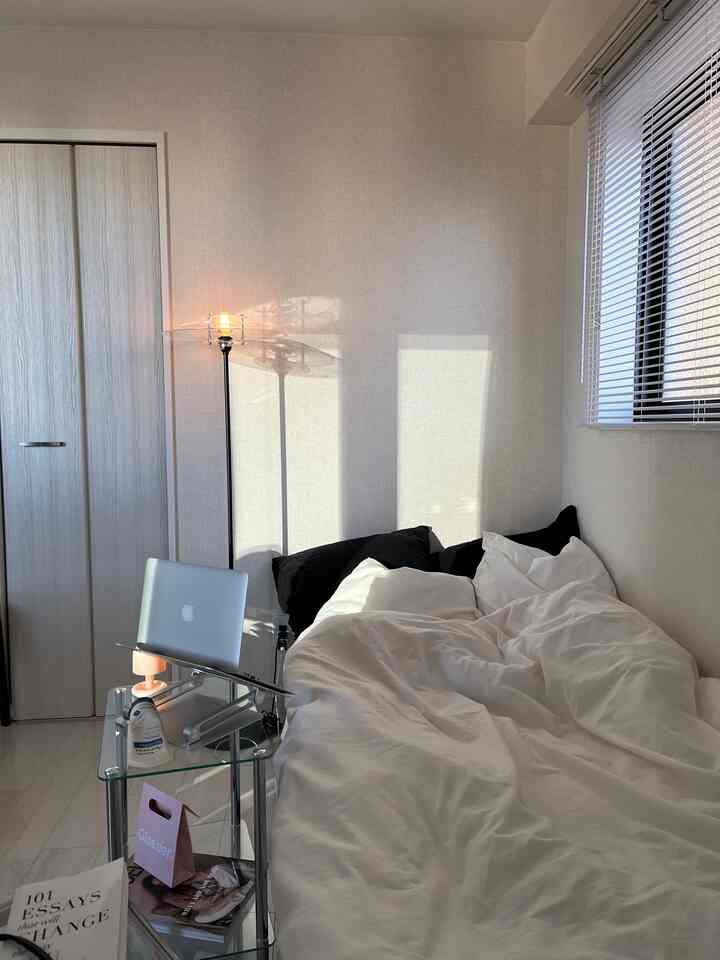 白を基調とした寝室、中央左にガラスのサイドテーブルとノートパソコン、右側にベッドとブラインドが配置された落ち着いた空間