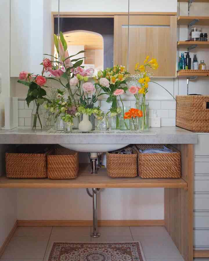 Natural toned bathroom sink area featuring multiple vases with flowers and rattan baskets under the countertop creating a cozy atmosphere