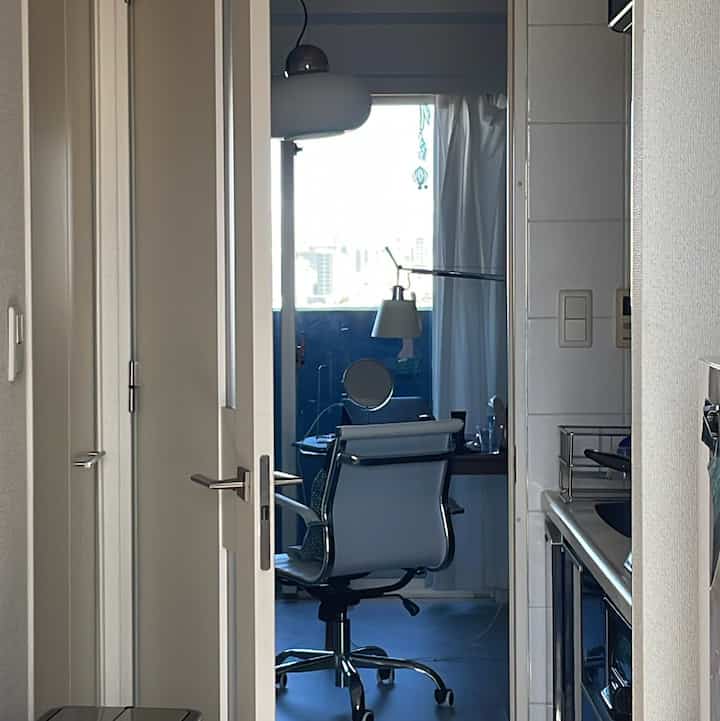 白と青を基調とした小さな玄関から繋がるホームオフィス、オフィスチェアと窓越しの街並みが印象的な落ち着いたインテリア