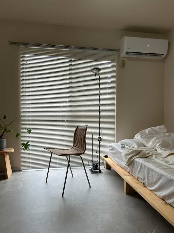 白と木目調の調和が取れた寝室、シンプルなスツールとフロアランプが配置された自然でインダストリアルな雰囲気のインテリア