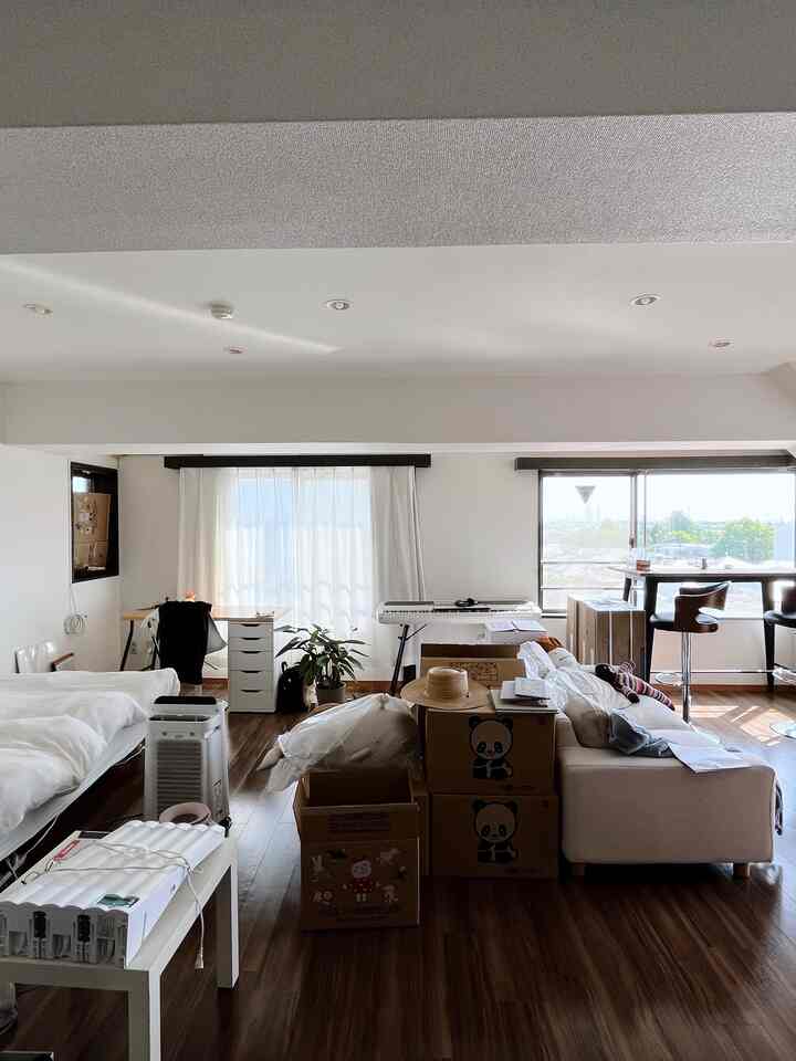 A narrow studio apartment in white and brown wood tones featuring a sofa bed, desk, dresser, and stacked boxes in a practical layout
